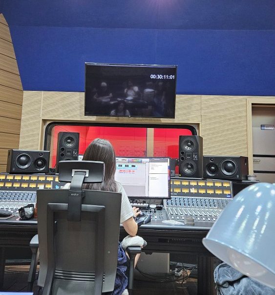 A sound engineer at a large mixing console with a film playing on the screen above.